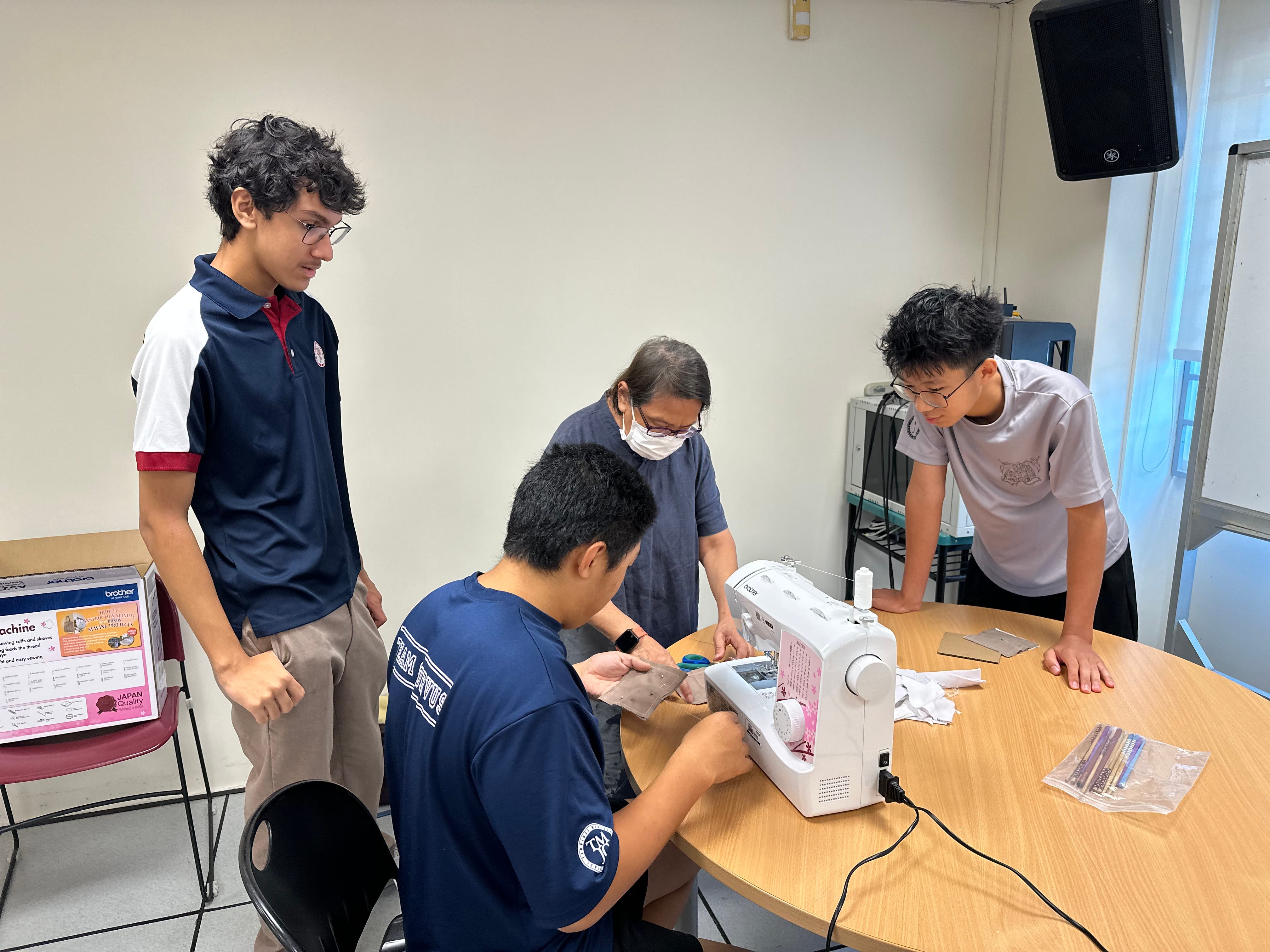 Four people — three students and an adult instructor wearing a mask — huddle around a Brother sewing machine on a round table, with one student operating the machine while the others observe.